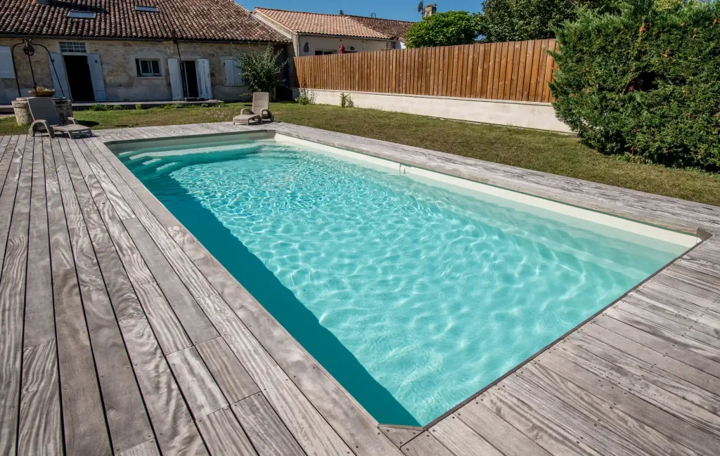 Piscine turquoise sur une grande terrasse en bois gris avec transats, maison en pierre ancienne et clôture en bois en arrière-plan