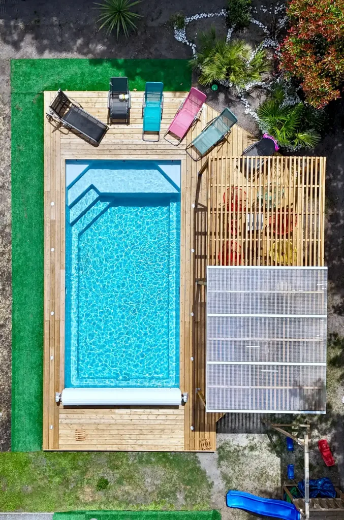 Vue aérienne d’une piscine rectangulaire turquoise sur terrasse en bois avec plusieurs transats colorés et gazon