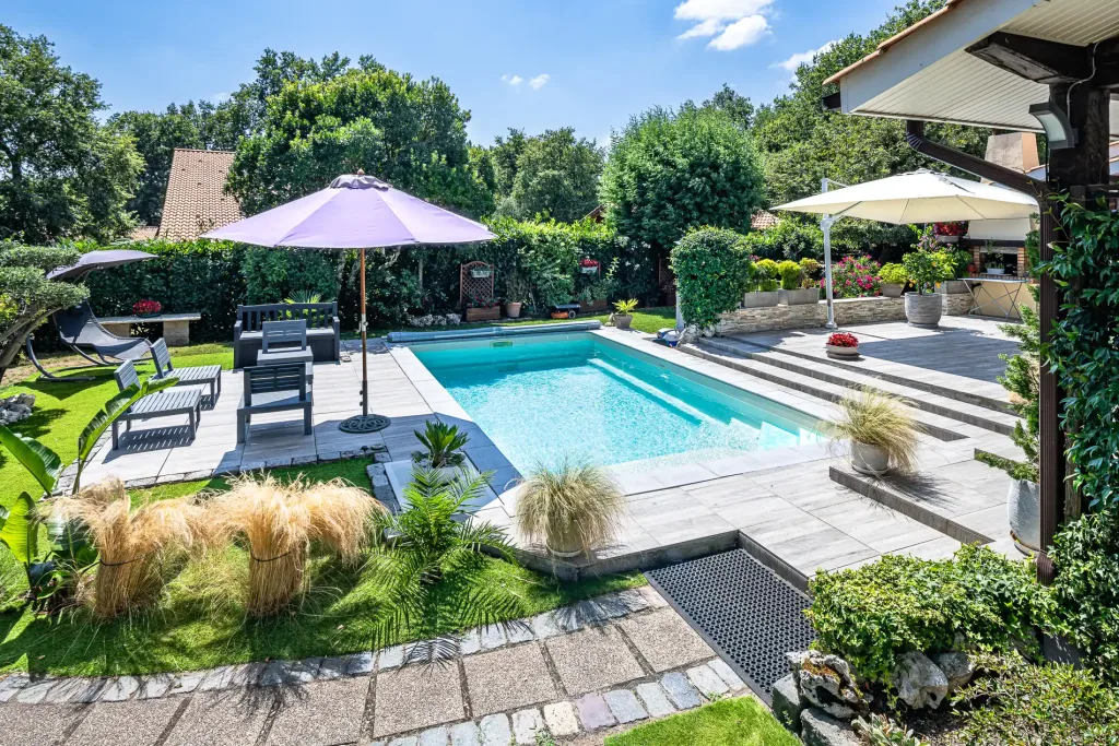 Piscine rectangulaire turquoise dans un grand jardin paysager avec terrasse en pierre, parasols et mobilier extérieur