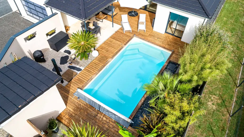 Vue aérienne d’une piscine rectangulaire turquoise entourée d’une grande terrasse en bois avec salon et transats