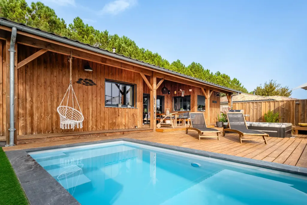 Piscine rectangulaire turquoise devant une maison en bois avec transats, jacuzzi et balançoire suspendue