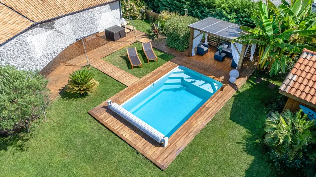 Vue aérienne d’une piscine rectangulaire turquoise sur grande terrasse en bois avec salon de jardin et pelouse
