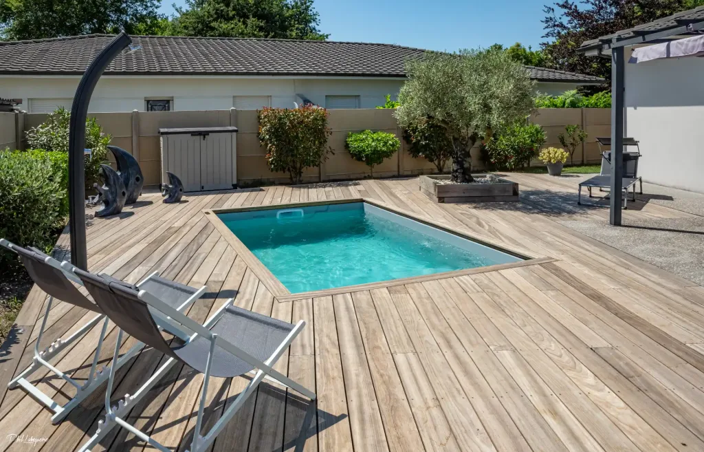 A small rectangular pool on a large wooden deck with lounge chairs and a garden