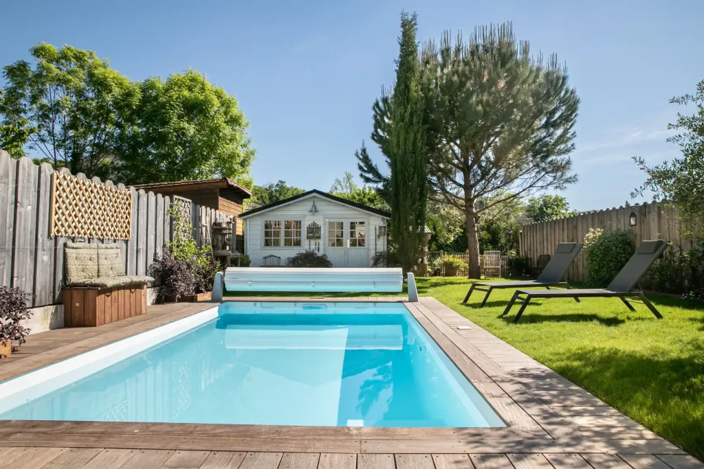 Piscine rectangulaire turquoise dans un jardin avec cabanon blanc, transats et clôture en bois