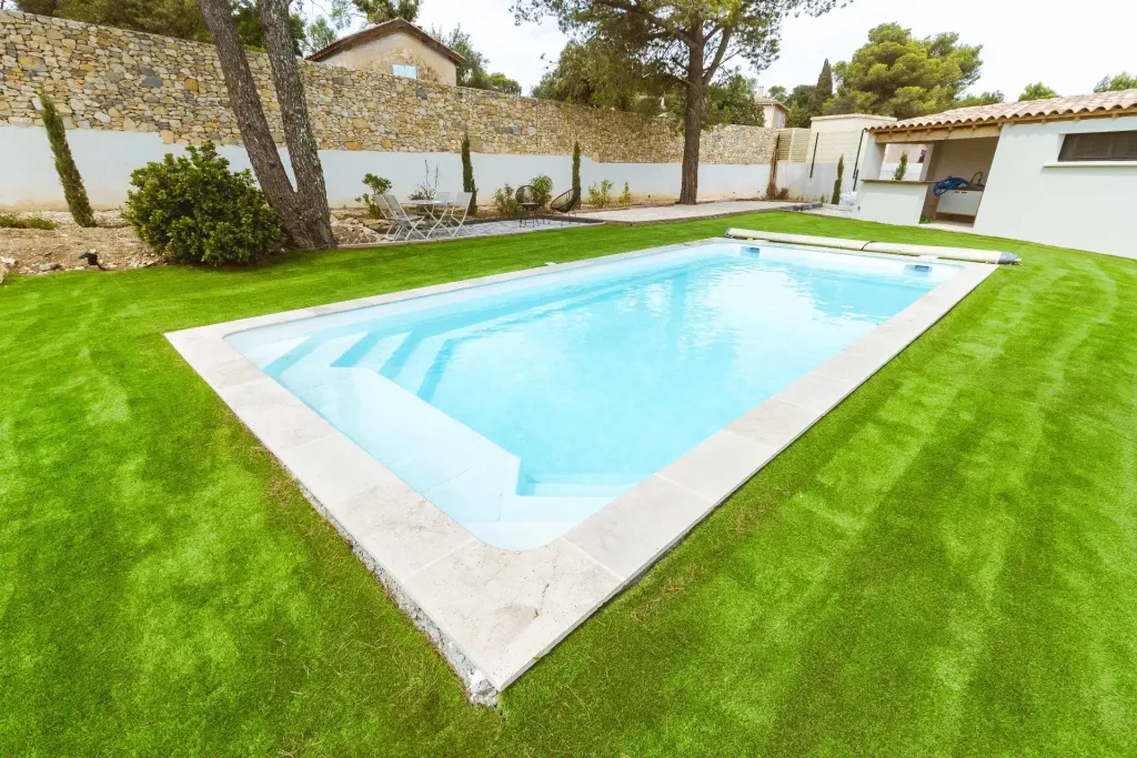 Piscine rectangulaire moderne entourée d'une grande pelouse verte, avec mur en pierre, arbres et espace détente en arrière-plan