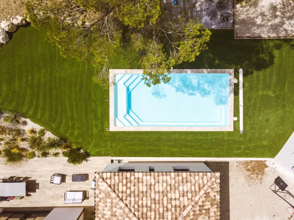 Vue aérienne d'une piscine rectangulaire turquoise dans un jardin avec pelouse synthétique verte, coin détente avec table et chaises, et toiture en tuiles en premier plan