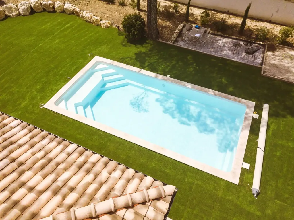 Vue aérienne d'une piscine rectangulaire bleue sur une pelouse verdoyante, avec terrasse en bois, toiture en tuiles et arbres environnants