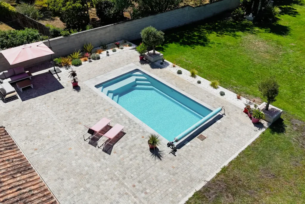 Vue aérienne d'une piscine moderne entourée d'une terrasse en carrelage clair, pelouse et coin repas avec parasol vert.