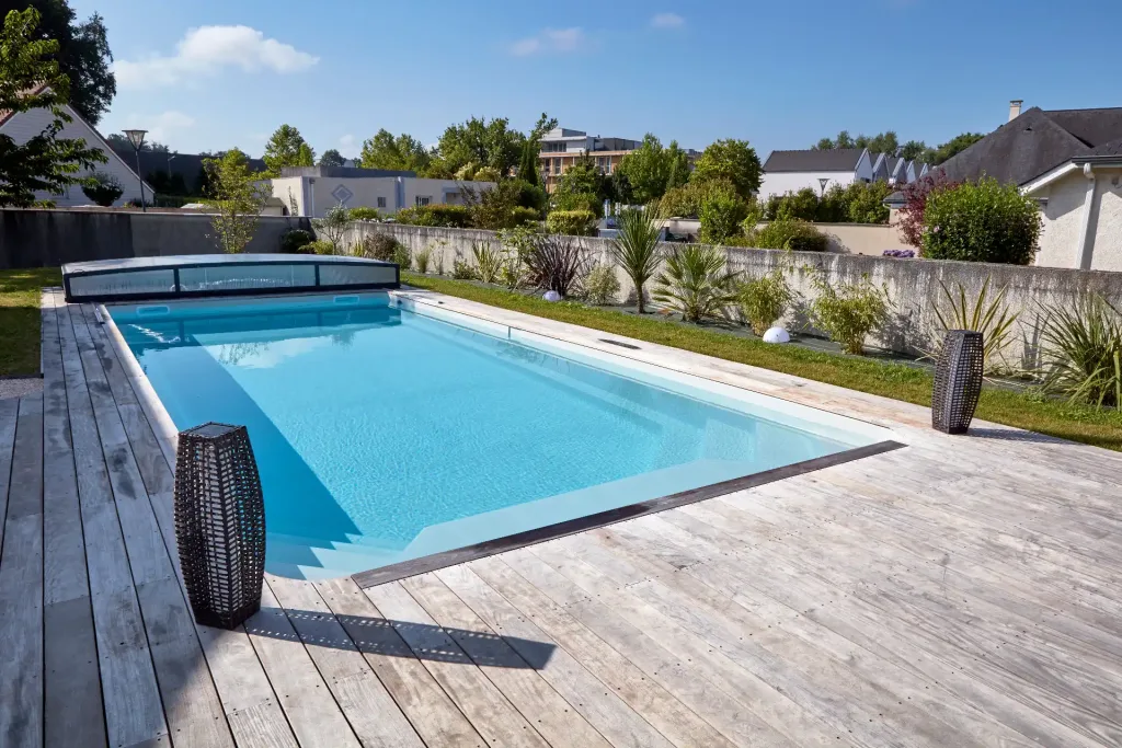 Piscine turquoise rectangulaire sur une grande terrasse en bois gris clair, avec lanternes décoratives noires, jardin paysager et maisons en arrière-plan sous un ciel bleu