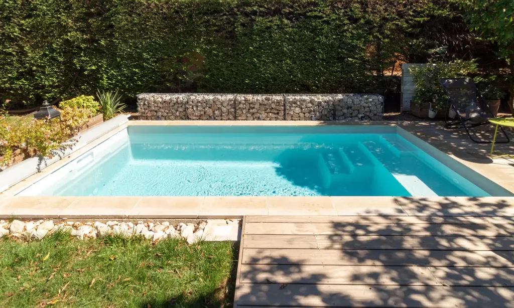 Mini-piscine rectangulaire turquoise entourée de haie végétale dense et terrasse en bois