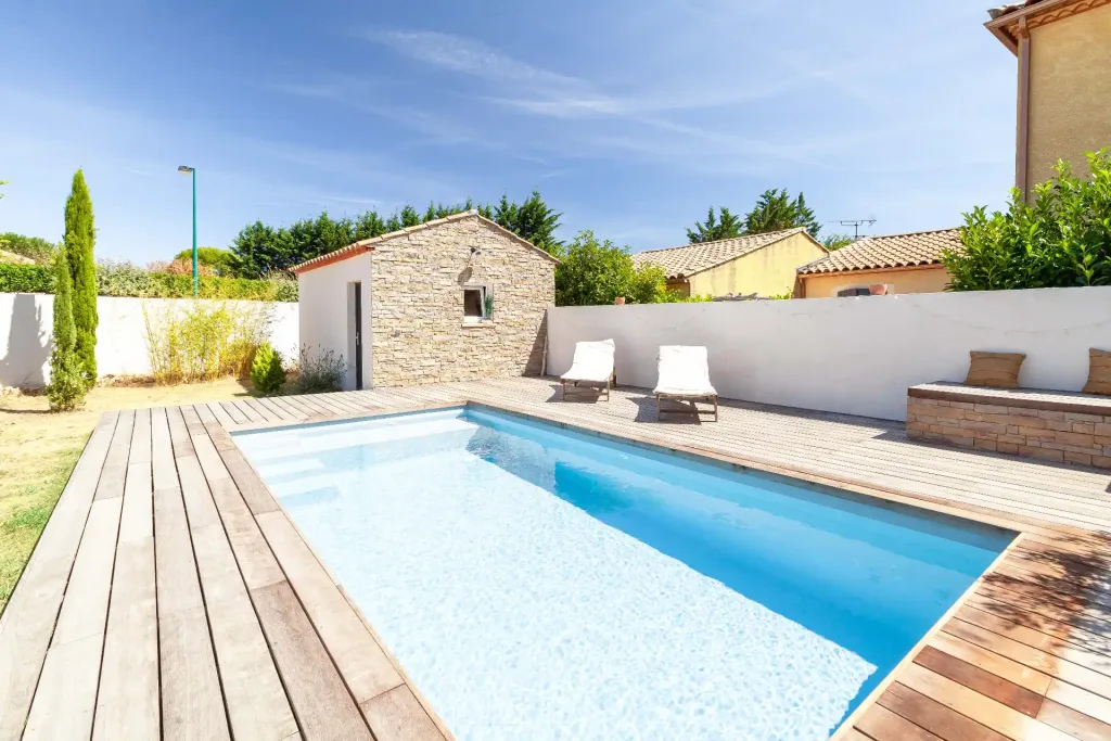 Piscine rectangulaire bleue entourée d’une grande terrasse en bois avec transats et mur blanc