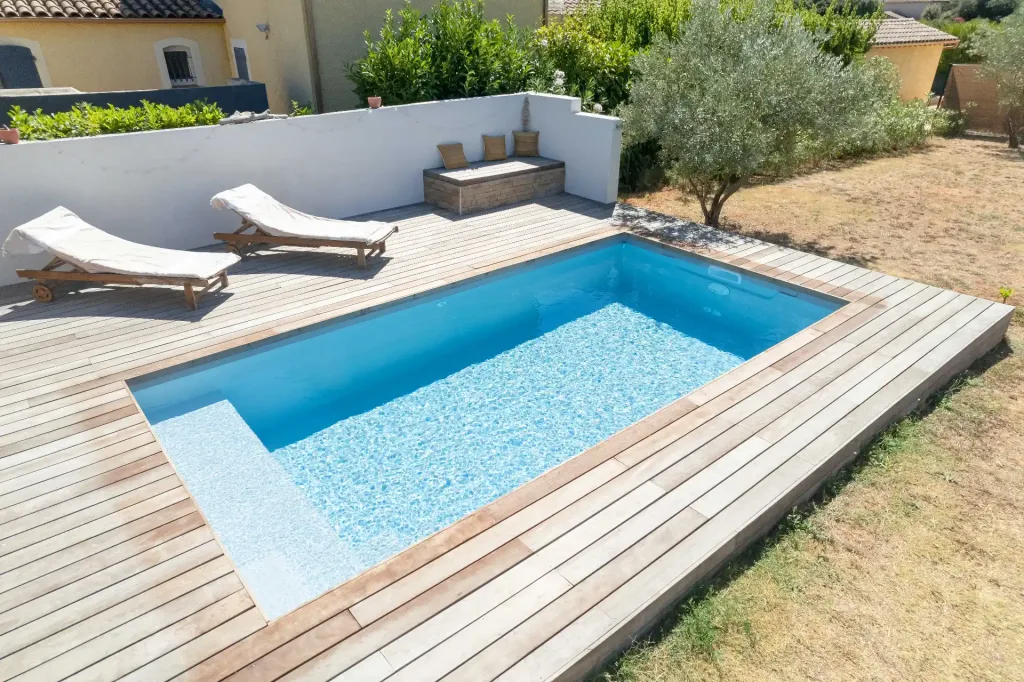 Piscine rectangulaire turquoise sur terrasse en bois avec transats blancs et petite maison en pierre