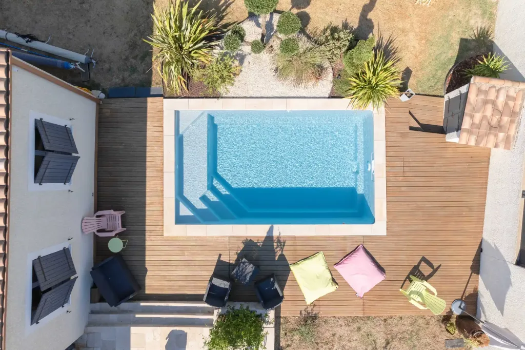Vue aérienne d’une piscine rectangulaire turquoise avec escalier sur terrasse en bois entourée de plantes