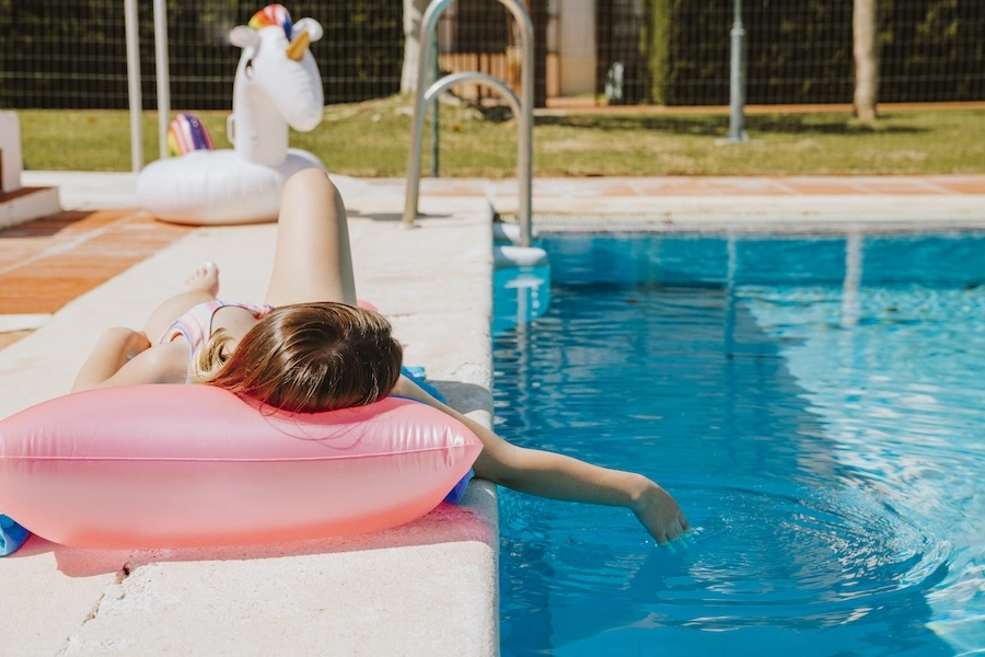Jeune femme allongée au bord d'une piscine extérieure, avec une clôture visible en arrière-plan, illustrant l'importance des distances réglementaires entre une piscine et la limite de propriété