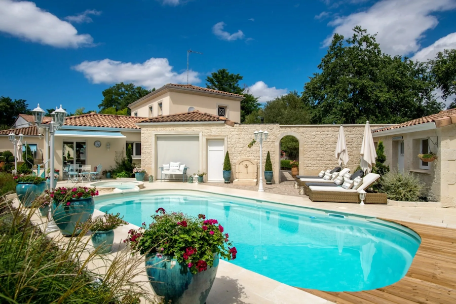 Piscine coque de forme ovale intégrée dans le jardin d'une maison individuelle provençale, entourée d'une terrasse en bois et de margelles en pierre beige