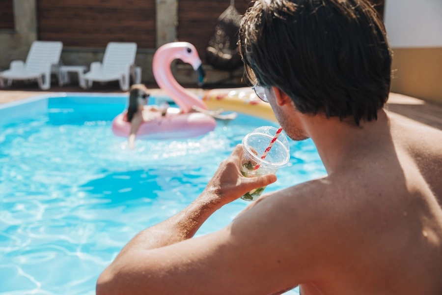 Propriétaire profitant de sa piscine en été avec un verre à la main, bouées flamant rose en arrière-plan