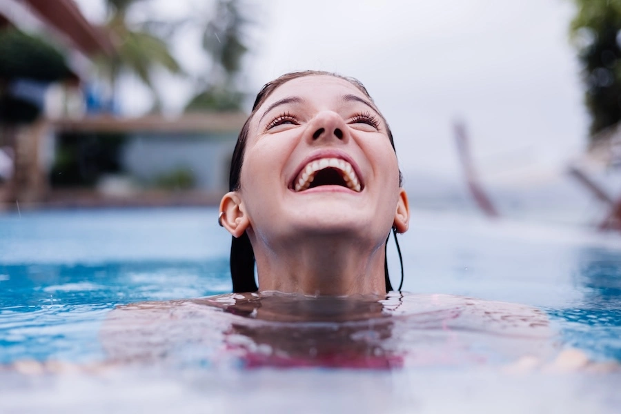 L&auml;chelnde junge Frau, die aus dem Wasser eines Schwimmbeckens auftaucht, ein Symbol f&uuml;r Vergn&uuml;gen und Entspannung am Beckenrand